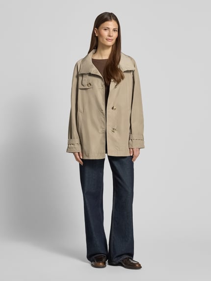 Frauenmodel trägt Trenchcoat mit Stehkragen und Ärmelriegeln von Fuchs Schmitt in Beige, lockerer Schnitt, Eingrifftaschen, Ganzkörperansicht von vorne.
