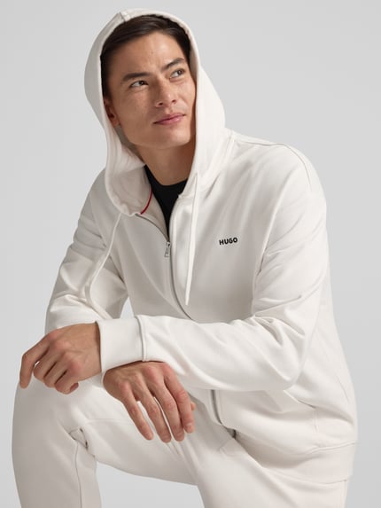 Männermodel trägt Relaxed Fit Sweatjacke aus reiner Baumwolle Modell 'DAPOZIP' von HUGO, weiß, Kapuze, Label Print, Nahaufnahme Oberkörper