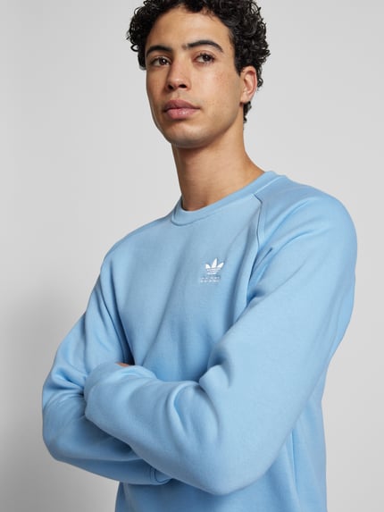Männermodel trägt Sweatshirt mit Label-Stitching von adidas Originals in Hellblau, Baumwoll-Mix, lockerer Schnitt, Nahaufnahme Oberkörper.