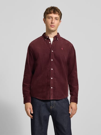 Męski model nosi Regular Fit Freizeithemd mit Logo-Stitching Modell 'MADISON' von Carhartt Work In Progress z bawełny, kołnierzyk typu button down, listwa guzikowa, sportowe mankiety, wyhaftowane logo, zaokrąglony dół, długość tyłu w rozmiarze M: 75 cm, szerokość w rozmiarze M: 47 cm, długość rękawów w rozmiarze M: 65 cm, kolor bordowy, całość o wyraźnej fakturze sztruksu. Ganzkörperansicht von vorne.