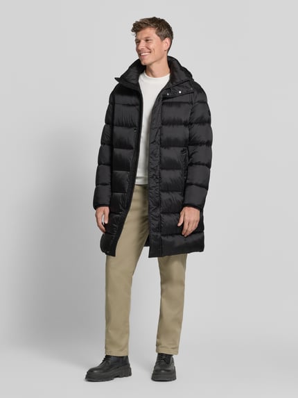 Männermodel trägt schwarze Pufferjacke mit Kapuze Modell 'JC-315Norman' von JOOP!, Longline, unifarben, Logo-Patch, Ganzkörperansicht von vorne.