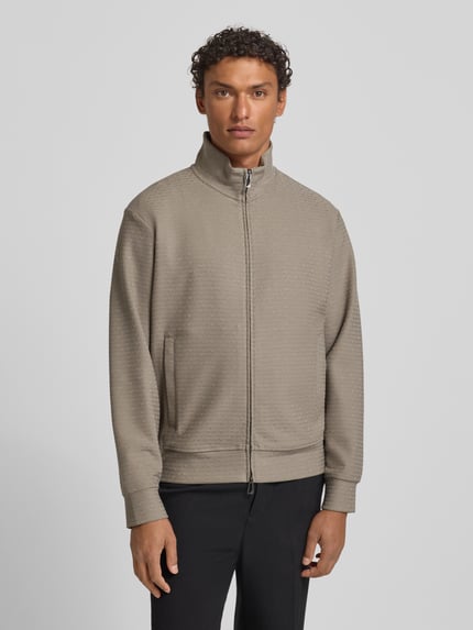 Männermodel trägt Sweatjacke mit Stehkragen von Emporio Armani in Beige, Baumwoll-Lyocell-Mix mit Stretch-Anteil, lockerer Schnitt, Reißverschluss, Eingrifftaschen, gerippte Abschlüsse, Rückenlänge 64 cm, Ärmellänge 60 cm, Ganzkörperansicht von vorne.