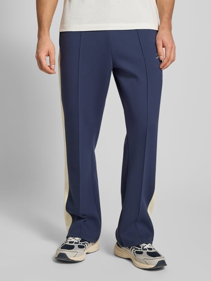 Männermodel trägt Sweatpants mit Längsnähten und Label-Print von KARL KANI in Blau mit cremefarbenen Seiten, Material-Mix, Nahaufnahme Unterkörper.