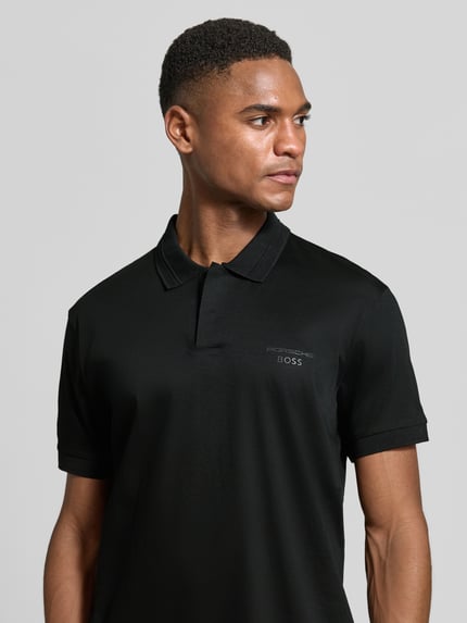 Männermodel trägt Regular Fit Poloshirt aus reiner Baumwolle Modell 'PS Spirit 70 Plk' von BOSS Green X PORSCHE in Schwarz, Nahaufnahme Oberkörper.