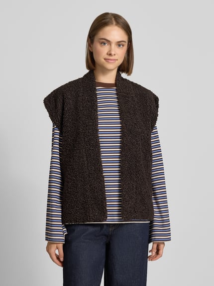 Modelka kobieca nosi czarną kamizelkę z imitacji futerka Regular Fit Weste in Teddy-Optik Modell 'NAYA' od ONLY na pasiastą bluzkę, widok całej sylwetki z przodu.