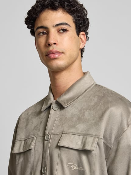 Męski model nosi Relaxed Fit Hemdjacke mit Brusttaschen und Label-Stitching od Pegador z weluru, kołnierzyk typu kent, listwa guzikowa, kieszenie na piersi, wyhaftowane logo, kolor beżowy, rozmiar M, długość tyłu 70 cm, zbliżenie na górną część ciała.