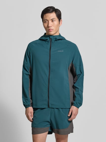 Męski model nosi Regular Fit Jacke mit Logo-Print Modell 'Division' od HALO, zielono-szara, lekka tkanina, z kapturem, widok z przodu, krój regular fit, elastyczny materiał, zamek, kieszenie, elastyczne mankiety, długość tyłu 67 cm, szerokość w rozmiarze M 40 cm ramion, 54 cm klatki piersiowej, rękaw 56 cm