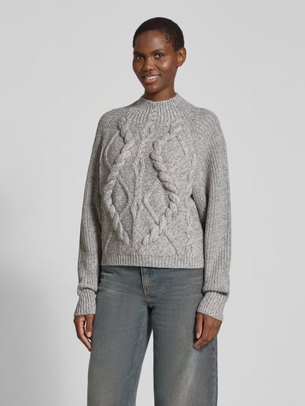 Frauenmodel trägt Pullover mit markantem Zopfmuster Modell 'Frescienne' von BOSS in grauer Melange-Strickoptik, hochgeschlossen, Ganzkörperansicht von vorne.
