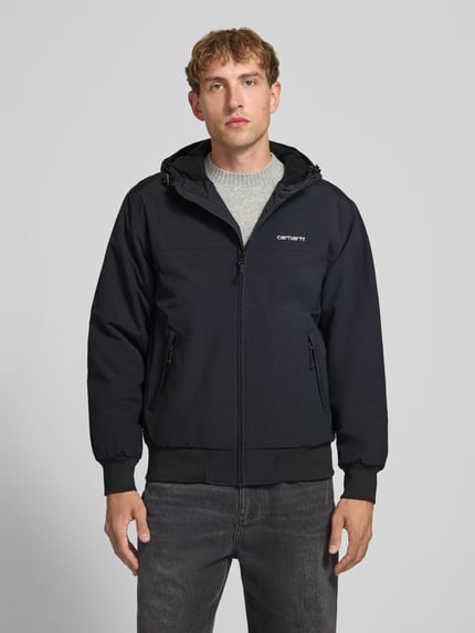 Männermodel trägt schwarze Funktionsjacke mit Logo-Stitching und Kapuze von Carhartt Work In Progress, Reißverschlusstaschen, gerippte Abschlüsse, Ganzkörperansicht von vorne.
