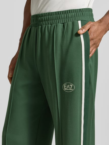 Männermodel trägt grüne Trackpants aus Viskose-Mix von EA7 Emporio Armani mit Galonstreifen und Label-Print, Nahaufnahme Oberkörper.