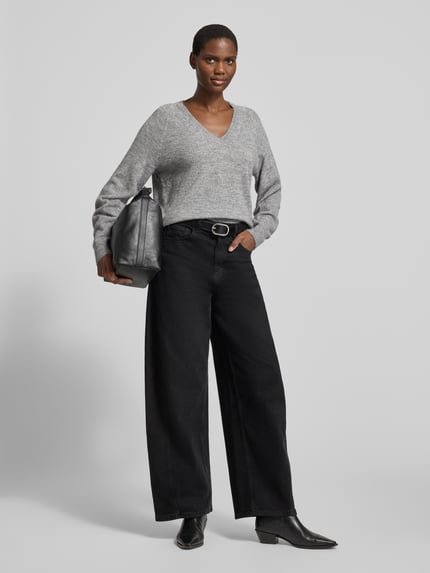 Frauenmodel trägt Pullover mit V-Ausschnitt VMTEAK von VERO MODA in grauer Melange-Strickoptik, schwarze weite Hose, Ganzkörperansicht von vorne.