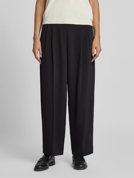Kobieta nosi spodnie 'Tapered Fit Hose mit Bundfalten' od COMMA, czarne, elastyczna mieszanka materiałów, krój tapered fit, zakładki w pasie, zapięcie na haftkę i zamek błyskawiczny, szerokość w rozmiarze 36: 48 cm, długość wewnętrzna nogawki: 70 cm, widok z przodu na całe ciało.