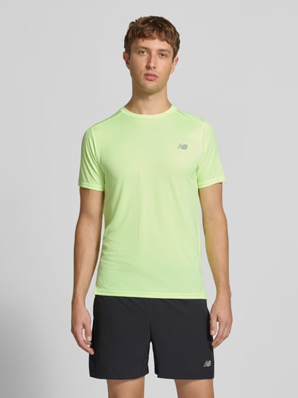 Männermodel trägt Funktionsshirt mit Rundhalsausschnitt und Logo von New Balance in Hellgelb, kombiniert mit schwarzen Shorts, Ganzkörperansicht von vorne. Rückenlänge bei Größe M: 69 cm, Regular Fit.