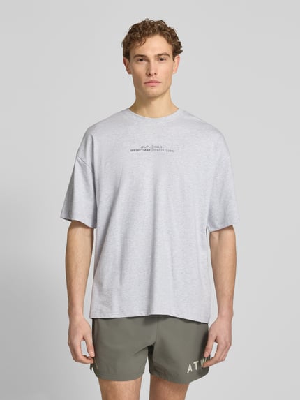Männermodel trägt T-Shirt mit Label-Print und Rundhalsausschnitt von HALO in Hellgrau, Boxy Fit, reine Baumwolle, Ganzkörperansicht von vorne.