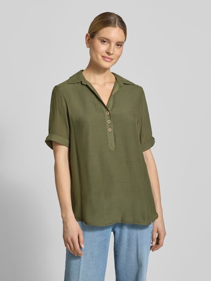 Vrouwenmodel draagt Relaxed Fit blouse uit viscosemix met platte kraag, knoopsluiting, halflange mouwen, semi-transparante look, verlengde achterkant van Apricot, kleur olijfgroen, ruglengte maat S 82 cm, mouwlengte 28 cm. Vooraanzicht halflijfopname.