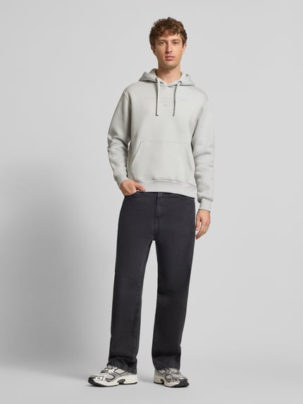 Männermodel trägt Relaxed Fit Hoodie aus Baumwoll-Mix mit Label-Detail von Alpha Industries in Hellgrau, Känguru-Tasche, Ganzkörperansicht von vorne.