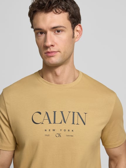 Männermodel trägt T-Shirt mit Label-Print und Rundhalsausschnitt von CK Calvin Klein in Beige, Reine Baumwolle, Regular-Fit, Nahaufnahme Oberkörper.