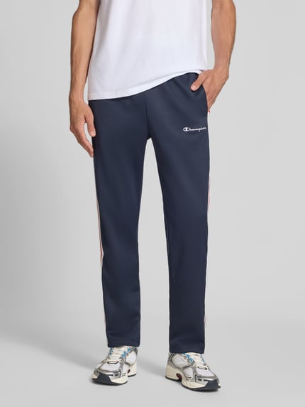 Männermodel trägt Sweatpants mit Bordüre von Champion in Dunkelblau, Material-Mix, gerader Schnitt, Seitentaschen, Nahaufnahme Unterkörper.