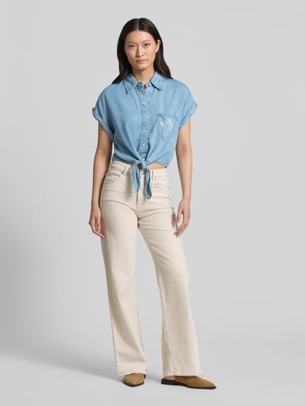 Frauenmodel trägt Jeansbluse mit Brusttasche Modell 'AMBRA' von Guess aus Lyocell in Hellblau, kombiniert mit heller Hose, Ganzkörperansicht von vorne.