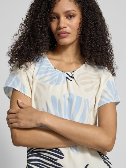 Modelka nosi Loose Fit Blusenshirt mit Allover-Muster od s.Oliver BLACK LABEL, efekt: wzór na całej powierzchni, okrągły dekolt, kontrafałda, krótki rękaw, wykończenie: lekko zaokrąglone, wykończenie materiału: szyfon, jasne kolory, detale w odcieniach niebieskiego i czerni, widoczna struktura materiału; zbliżenie na górną część ciała, widok z przodu.