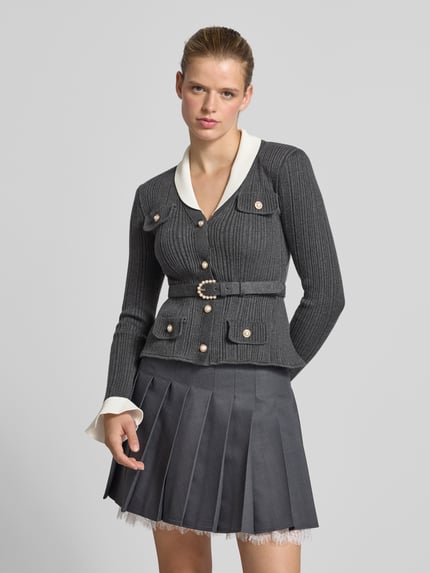 Modelka nosi kardigan mit Viskose-Woll-Anteil von Self-Portrait, Mieszanka materiałów, krój slim fit, imitacja dzianiny, kieszenie z patką, z długim rękawem, długość tyłu w rozmiarze S: 57 cm, odcień szarości z białym kołnierzem, przód z ozdobnym paskiem, szara plisowana spódnica, gładka tekstura, widok całej sylwetki z przodu.