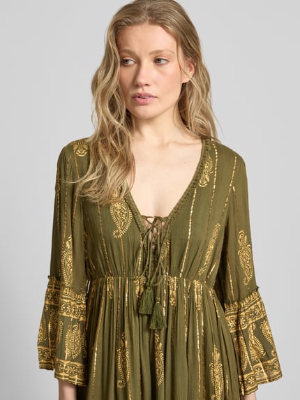 Frauenmodel trägt Minikleid aus Viskose-Mix mit Schnürung von Apricot in Oliv mit goldenen Mustern, Trompetenärmel, Nahaufnahme Oberkörper. Rückenlänge bei Größe S: 83 cm, Brustweite: 35 cm, Ärmellänge: 50 cm, A-Linie, V-Ausschnitt.