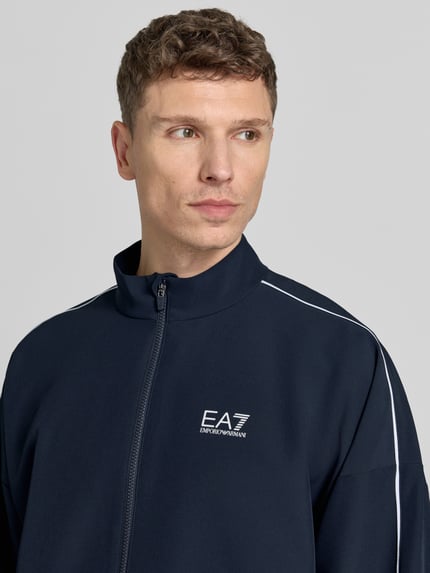 Męski model nosi Bomberjacke mit Stehkragen und Logo-Print od EA7 Emporio Armani, kolor granatowy, logo na piersi, zbliżenie na górną część ciała. Szerokość w rozmiarze M: 66 cm, długość tyłu: 71 cm, szerokość ramion: 75 cm, długość rękawa: 54 cm, tkanina VENTUS7.