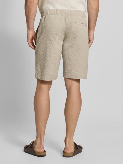 Männermodel trägt szorty z fakturowanym wzorem z wpuszczanymi kieszeniami, kieszenią z tyłu, elastycznym pasem i tunelem Shorts mit elastischem Bund und Eingrifftaschen von MCNEAL, krój regular fit, kolor jasnobeżowy, szerokość w rozmiarze M: 44 cm, długość wewnętrzna nogawki: 26 cm, zdjęcie całej sylwetki z tyłu