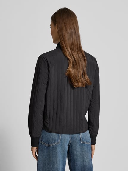 Frauenmodel trägt czarną kurtkę z pikowanymi przeszyciami Jacke mit Stehkragen und Eingrifftaschen von Scotch & Soda, krój regular fit, stójka, zamek błyskawiczny, wpuszczane kieszenie, detal z logo, długość tyłu 57 cm, szerokość ramion 44 cm, szerokość klatki 50 cm, długość rękawów 63 cm, ganzkörperansicht rückansicht.