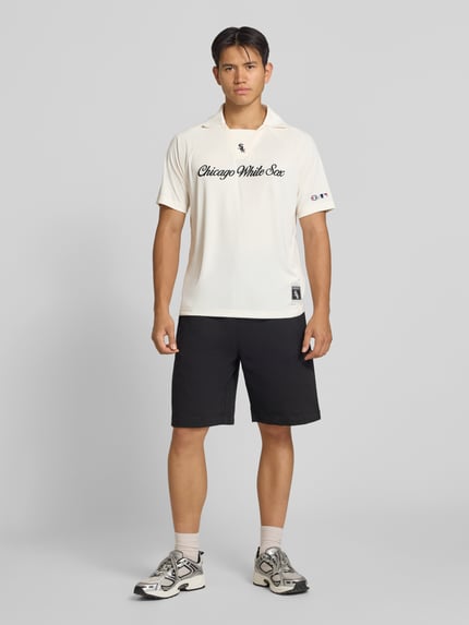 Männermodel trägt Relaxed Fit Poloshirt mit Logo-Stitching von Champion in Creme, schwarze Shorts, weiße Sneakers, Ganzkörperansicht von vorne. Herren Poloshirt mit Polokragen, kurzärmelig, Rückenlänge bei Größe M 70 cm.