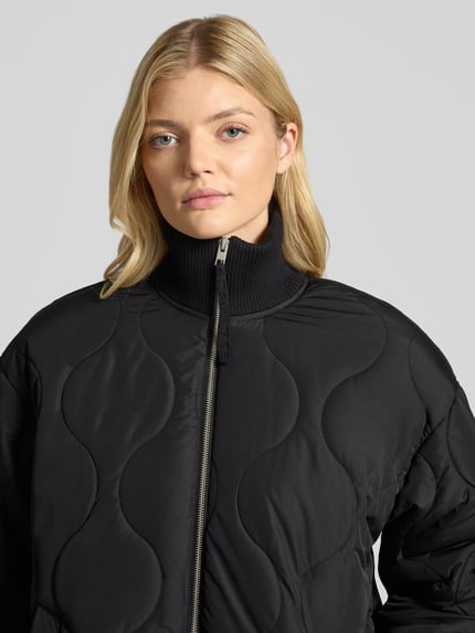 Kobiece model prezentuje Steppmantel mit Zweiwege-Reißverschluss Modell 'SERENA' von JJXX, czarny, krój regular fit, stójka, tkanina pikowana, szerokość w rozmiarze S: 59 cm, długość tyłu w rozmiarze S: 110 cm, szerokość ramion w rozmiarze S: 55 cm, długość rękawa w rozmiarze S: 54 cm, zbliżenie na górną część ciała.
