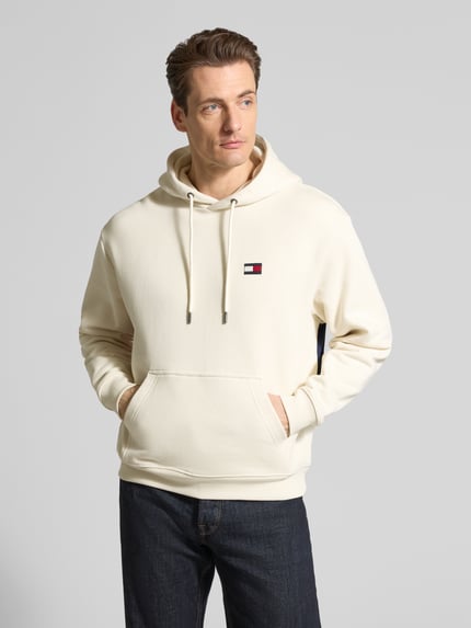 Mannenmodel draagt Regular Fit Hoodie aus reiner Baumwolle van Tommy Hilfiger in effen crème, logodetail voor, kangoeroezak, sweatstof, pure katoen, regular fit, lange mouw, geribde zoom, vooraanzicht bovenlichaam.