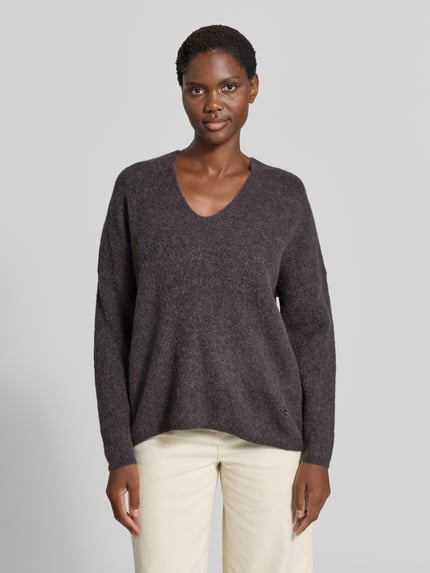 Kobieta nosi sweter z dzianiny z mieszanki wełny z domieszką alpaki, krój regular fit, dekolt w serek, model 'THORA' od MOS MOSH, długość tyłu w rozmiarze S: 82 cm, długość rękawów w rozmiarze S: 46 cm, ciemnoszary kolor, miękka tekstura, widoczne detale splotu, widok całej sylwetki z przodu.