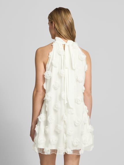 Vrouwenmodel draagt Minikleid met florale Applikationen van Gina Tricot in wit, loose fit, halter, druppelvormige hals, ruglengte maat S 83 cm, borstwijdte maat S 57 cm, achterkant volledig zichtbaar, volledige lichaamsopname rugaanzicht.