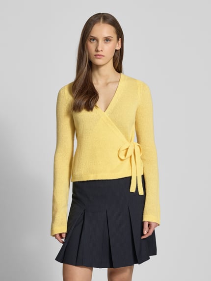 Modelka nosi Strick-Cardigan in Wickeloptik von REVIEW, krój regular fit, dekolt w serek, długie rękawy, kokarda, długość tyłu 50 cm, długość rękawów 66 cm, kolor żółty, gładka tekstura. Ganzkörperansicht von vorne.