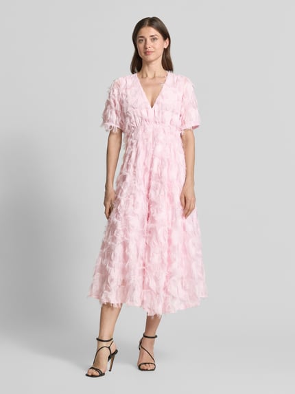 Frauenmodel trägt Cocktailkleid mit Fransen Modell 'PAZYLLA' von YAS in zartem Rosa, A-Linie Midikleid, V-Ausschnitt, Ganzkörperansicht von vorne.