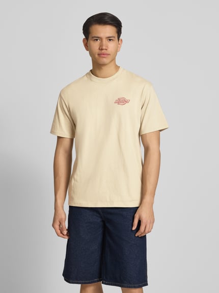 Męski model nosi T-Shirt mit Logo Modell 'MILLVILLE' von Dickies z bawełny, krój relaxed fit, okrągły dekolt, długość tyłu w rozmiarze M: 70 cm, widok całej sylwetki z przodu.