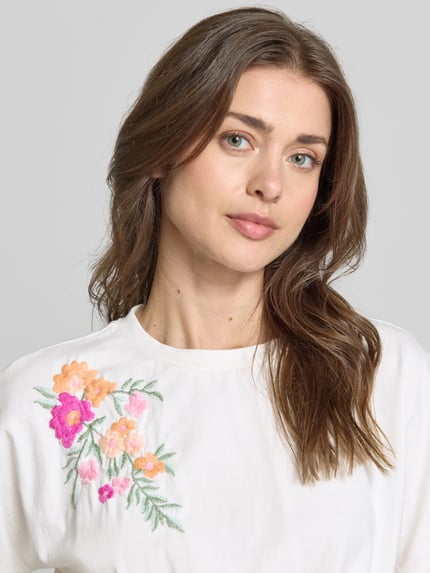 Modelka kobieca ma na sobie Regular Fit T-Shirt mit floralem Stitching Modell 'Fabian' od YAS, czysta bawełna, białe kolory z haftowanym kwiatowym wzorem, krój regular fit, ozdobny węzeł, długość pleców w rozmiarze S: 47 cm, zbliżenie na górną część ciała.