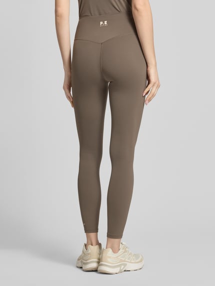Frauenmodel trägt Leggings mit elastischem Bund und Logo von P.E Nation in Braun, Material-Mix mit Stretch-Anteil, körpernahe Passform, High-Rise, Logo, Ganzkörperansicht Rückansicht.