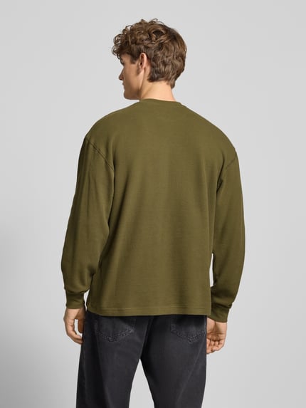 Männermodel trägt Longsleeve mit Label-Stitching von KARL KANI in Olivgrün aus reiner Baumwolle, lockerer Schnitt, Rückenlänge 68 cm, Ganzkörperansicht Rückansicht.