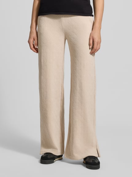 Vrouwenmodel draagt Wide Leg Sweatpants met Logo-Muster model 'LAUDOMIA' van GUESS Activewear, beige, elastische tailleband, materiaal-mix met stretch, logo patroon, binnenbeenlengte maat S 83 cm, volledig lichaam van voren.