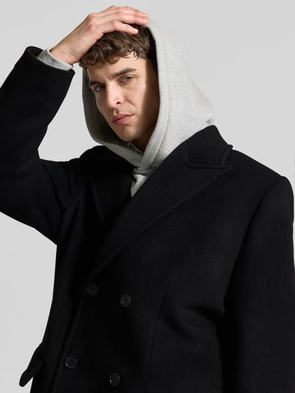 Męski model nosi Mantel aus Baumwoll-Mix od Filippa K, czarny, podwójny rząd guzików, patki na kieszeniach, prosta forma, długi rękaw, długość tyłu w rozmiarze 50: 135 cm, szerokość w rozmiarze 50: 48 cm, długość rękawa w rozmiarze 50: 67 cm, zbliżenie na górną część ciała z przodu.