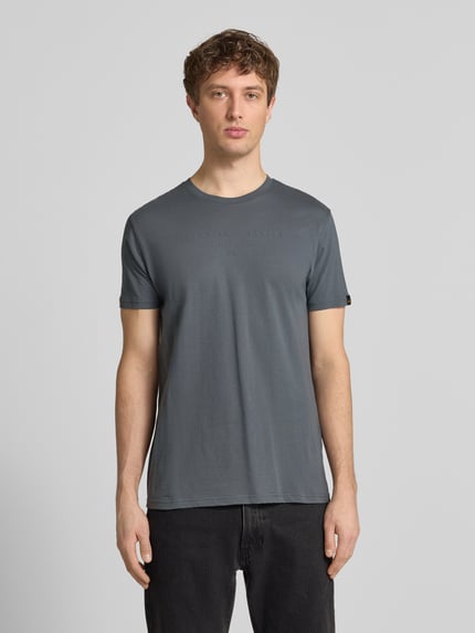 Męski model nosi T-Shirt aus Baumwolle von Alpha Industries w kolorze szarym, prosty krój, okrągły dekolt, detale z logo, długość tyłu w rozmiarze M: 68 cm, widok całej sylwetki z przodu.