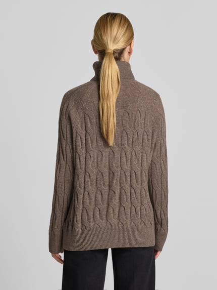 Männermodel trägt Relaxed Fit Rollkragenpullover aus reiner Wolle von Tommy Hilfiger in Braun mit Zopfmuster, Strukturstrick, Ganzkörperansicht Rückansicht. Pulower damski typu relaxed fit od Tommy Hilfiger z czystej wełny, brązowy, wzór warkoczowy, dzianina strukturalna, widok całej sylwetki od tyłu.