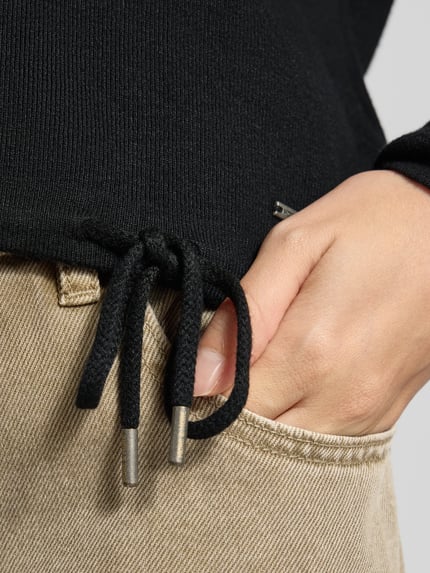 Frauenmodel trägt Strickpullover aus Viskose-Mix mit Rundhalsausschnitt von MORE & MORE, Detailaufnahme, Label-Detail, Tunnelzug, Nahaufnahme Hand, beige Jeans.