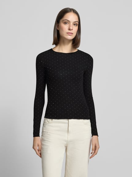 Modelka kobieca ma na sobie Langarmshirt mit schimmernden Details od VERO MODA, czarny kolor, połyskująca kropkowana faktura, długie rękawy, okrągły dekolt, damska góra, widok całej sylwetki z przodu.