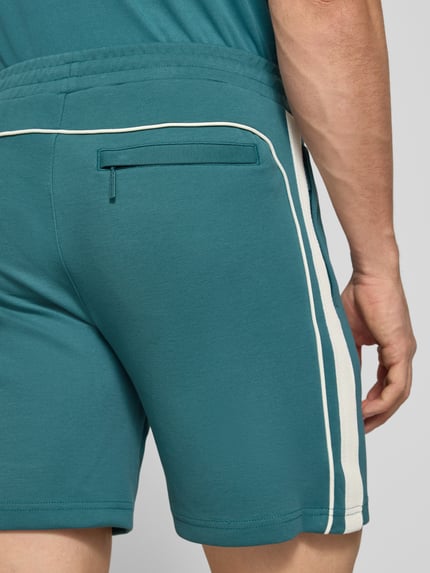 Männermodel trägt Shorts mit Logo-Stitching Modell 'EGEO' von SERGIO TACCHINI in Türkis mit weißen Streifen, Nahaufnahme Rückansicht. Baumwoll-Mix mit Stretch und elastischem Bund, lockerer Schnitt, Detailansicht der seitlichen Eingrifftaschen und Ziertasche.