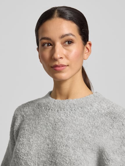 Modelka prezentuje Strickpullover mit gerippten Abschlüssen Modell 'ZOLLY' od American Vintage, jasnoszara faktura, półokrągły dekolt, luźny krój, miks wełny z alpaką, długość pleców 60 cm, długość rękawa 33 cm, zbliżenie górnej części ciała.