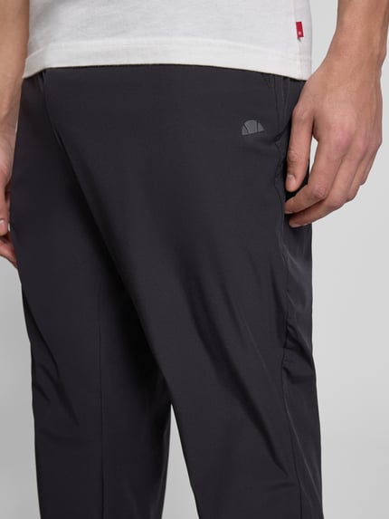 Mężczyzna nosi spodnie typu track pants z elastycznym pasem model 'EVARO' od Ellesse, mieszanka materiałów z dodatkiem streczu, krój regular fit, kolor czarny, długość wewnętrzna nogawki w rozmiarze M: 74 cm, zbliżenie na górną część nogawek z przodu.
