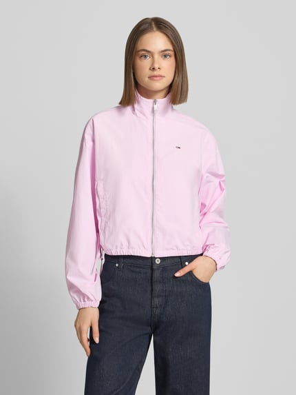 Frauenmodel trägt Relaxed Fit Blouson mit Taillenbändern von Tommy Jeans in Hellrosa, unifarben, Stehkragen, Logo-Stitching, Reißverschluss, lange Ärmel mit elastischen Abschlüssen, seitliche Eingrifftaschen mit Druckknopf, Ganzkörperansicht von vorne.